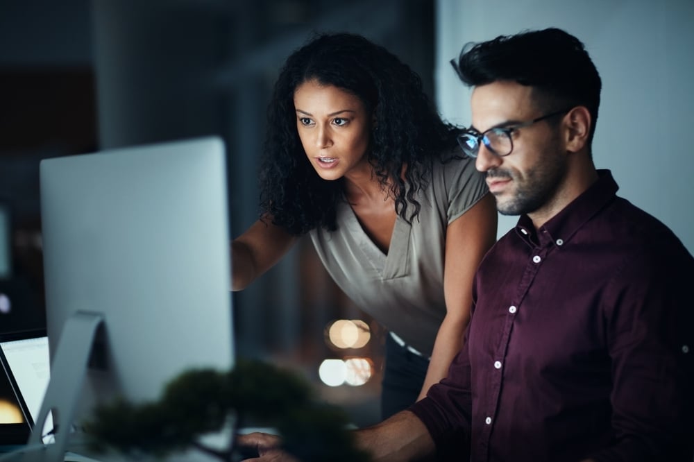 A woman and man collaborate at a computer late at night. She points at the screen with a focused expression.