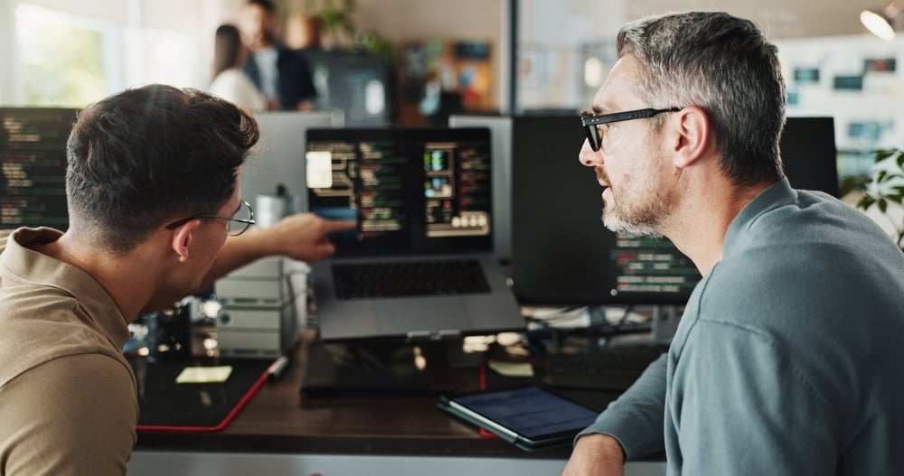 A developer points at code on a laptop, getting feedback from a senior colleague in a modern tech office.