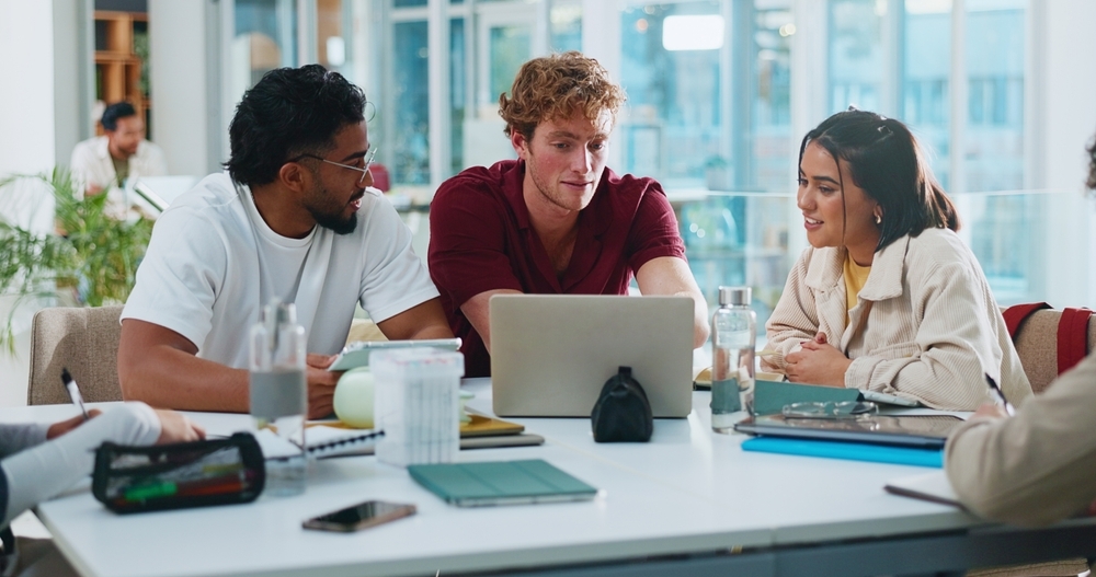 Three diverse students collaborate around a laptop at a table, studying together in a bright, modern room.