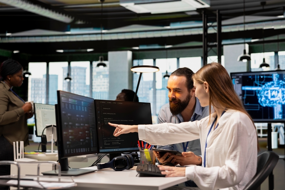 Two developers collaborating on AI software in a modern office. A woman points to code on a dual-screen display.