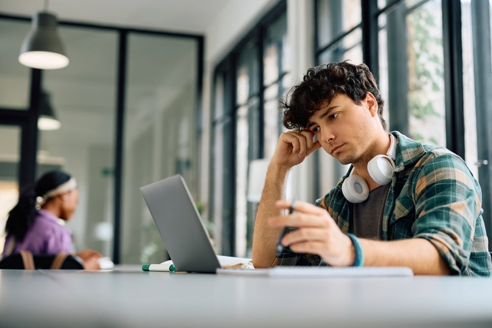 Focused young man in a plaid shirt looking at a laptop, resting his head on his hand with headphones around his neck.