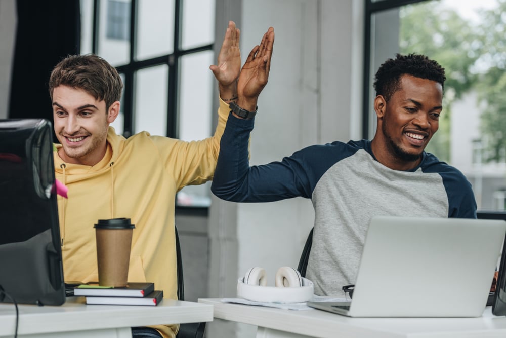 Two smiling male colleagues high-fiving across their desks while working on computers in an office.